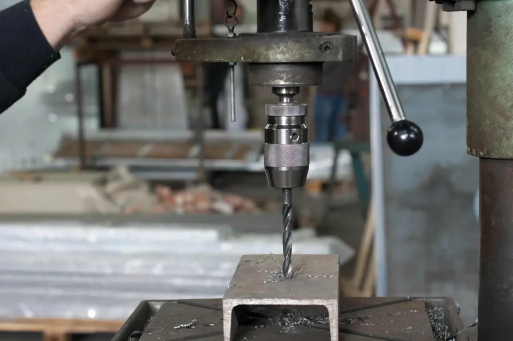 Un taladro con broca espiral taladra un orificio piloto en una viga metálica en I en un taller, preparando la pieza para el posterior roscado en la creación de roscas.