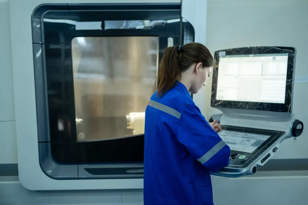 Female factory technician in blue work uniform operating digital touchscreen control panel of advanced CNC machining center to set production parameters.