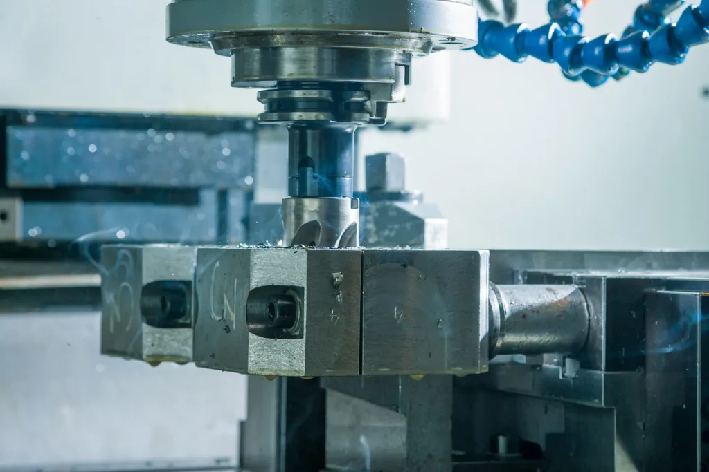 Vertical CNC machining center cuts steel block with rotating end mill, cooling hoses arranged alongside as metal shavings and faint smoke form during precision milling.