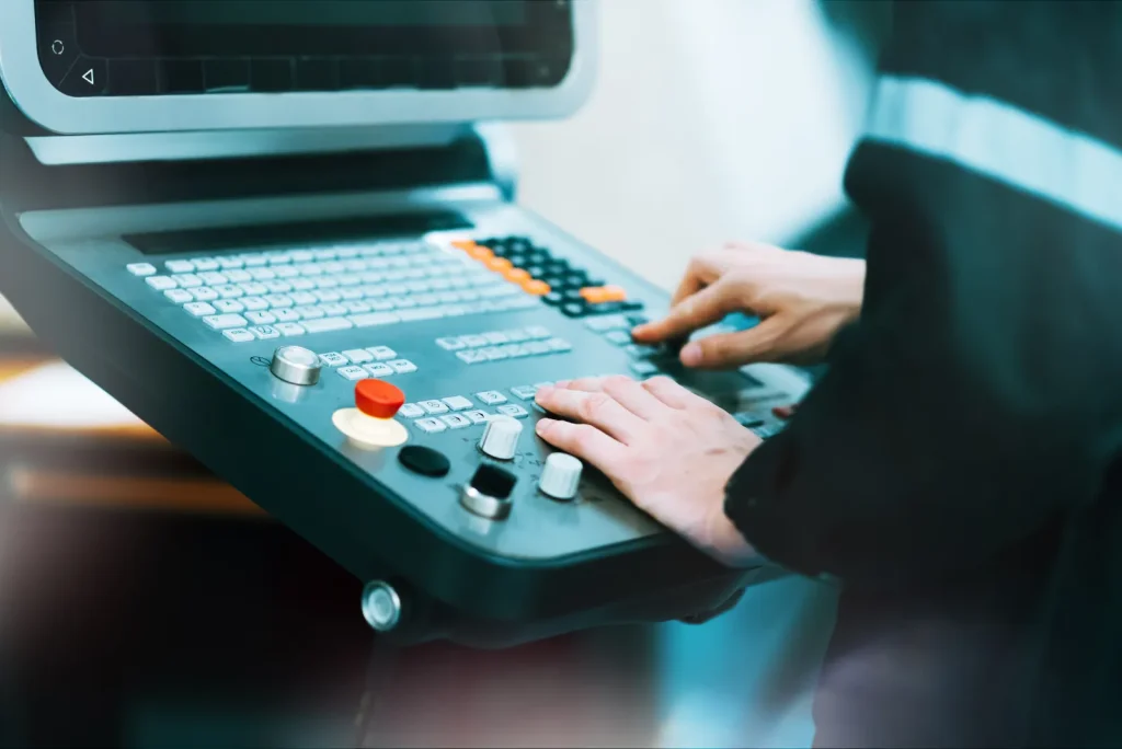 Factory worker’s hands operate physical CNC machine control panel with keyboards, rotary knobs and emergency stop button to input machining program parameters.