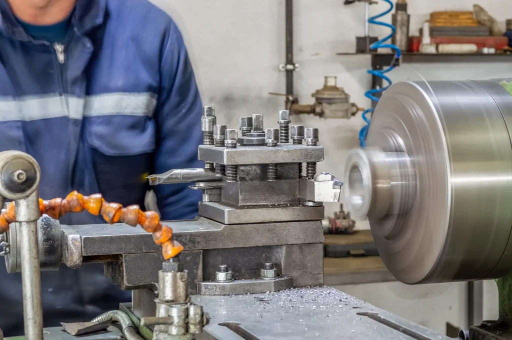 Technician in blue work uniform monitors a traditional metal lathe as the spindle spins rapidly, metal shavings pile on the machine bed after turning work.